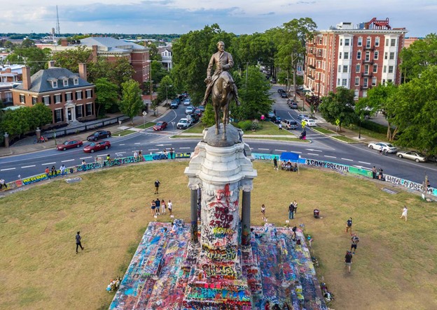 Aerial view from the front of the Robert E. Lee monument on Monument Avenue. The monument is covered with graffiti and there are people on the ground surrounding the monument.