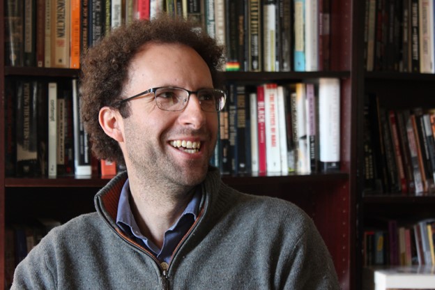 Dr. Adam Ewing wearing glasses and a gray quarter zip smiling and looking to his left in front of a bookshelf