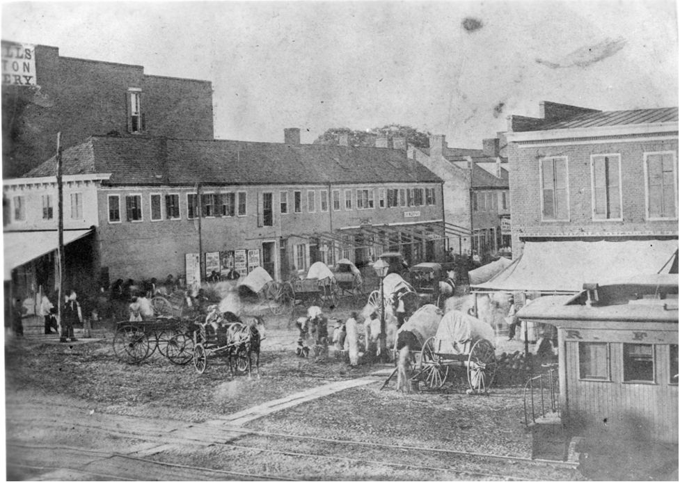 Black and white image of an intersection in Jackson Ward, horse-drawn covered wagons are shown parked along the streets and a railroad track with caboose runs through the intersection